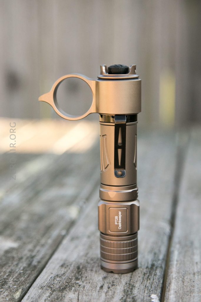 A bronze-colored flashlight with a circular ring attachment stands upright on a weathered wooden surface, with a blurred outdoor background.