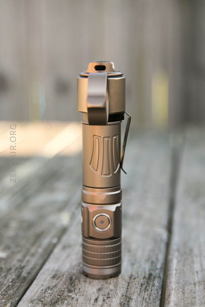 A metallic flashlight with a side clip stands upright on a weathered wooden surface, with a blurred outdoor background. The flashlight features a button and grooved grip on its lower section.