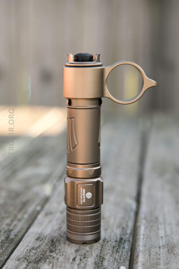 A bronze-colored tactical flashlight with a circular ring attachment stands upright on a wooden surface, shown in focus with a blurred wooden background.