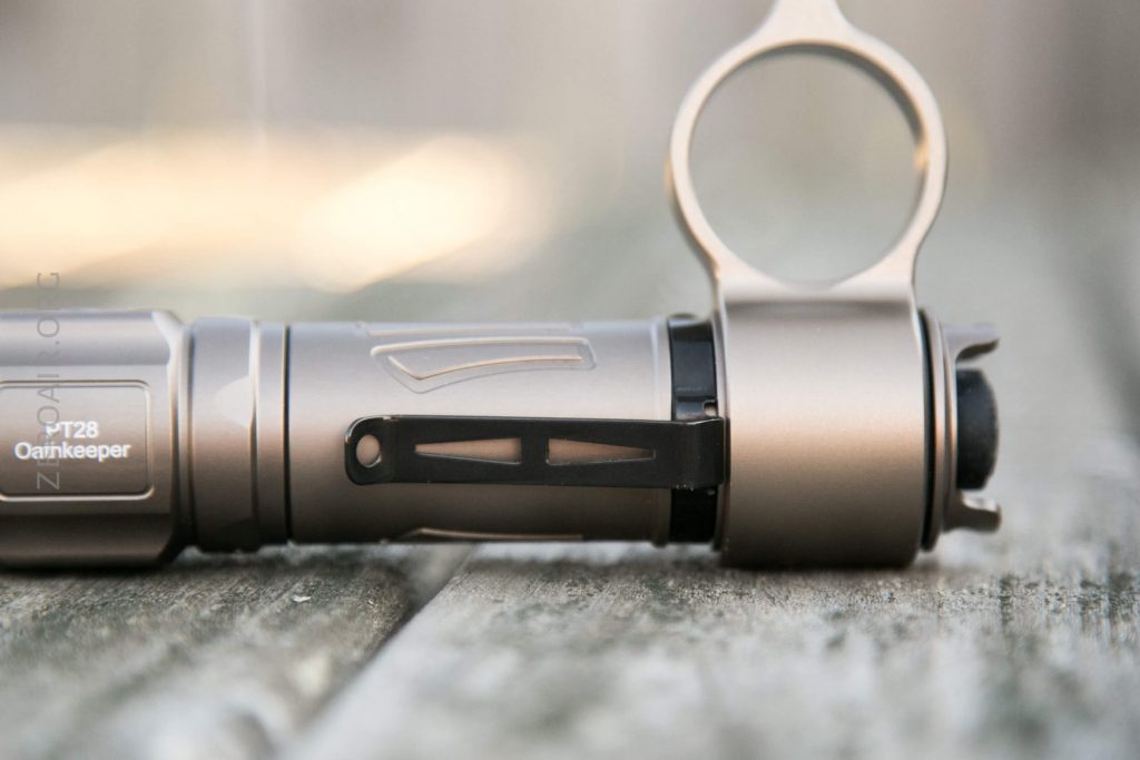 A close-up of a metallic flashlight with a side clip, labeled “T28 Oathkeeper,” lying on a wooden surface with a blurred background.