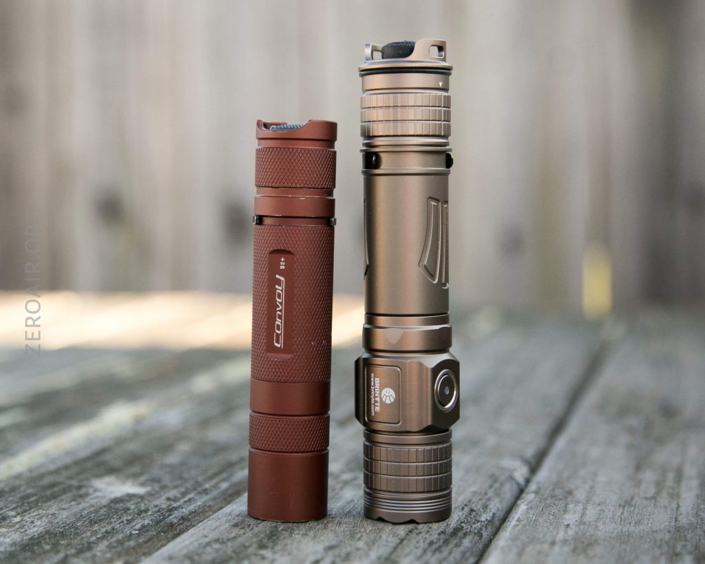 Two flashlights stand upright on a weathered wooden surface outdoors. The flashlight on the left is shorter and red, while the one on the right is taller, metallic, and bronze-colored. The background is blurred.