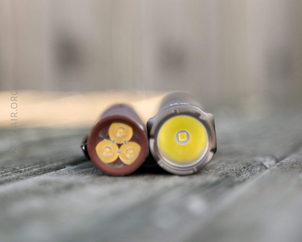 Two flashlight heads lying on their sides on a wooden surface, one with four yellow LED emitters and the other with a single yellow LED emitter, both out of focus in the background.