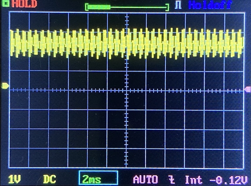 Oscilloscope screen showing a yellow rectangular waveform signal, with voltage set to 1V per division and a time base of 2ms per division. The display shows grid lines and measurement indicators.