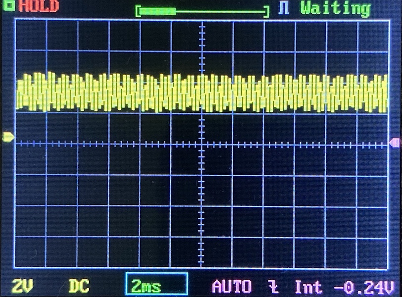 A digital oscilloscope screen displays a yellow waveform signal centered on the grid. The settings show 2V per division vertically and 2ms per division horizontally. The status reads Waiting in the top right corner.