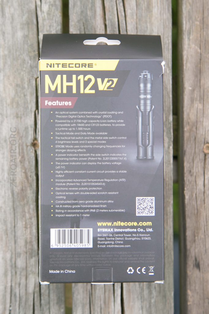 The back of a Nitecore MH12 V2 flashlight box is shown. The packaging lists features, technical specifications, a product image, company information, website, and a QR code, all on a wooden surface.