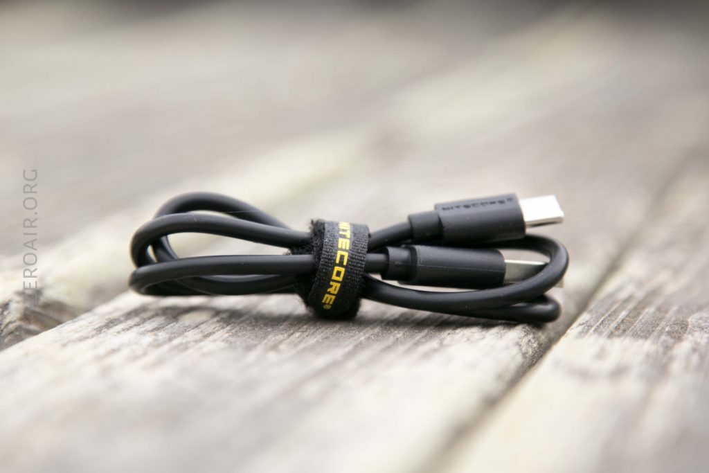 A coiled black USB-C cable secured with a black and yellow Nitecore-branded strap rests on a weathered wooden surface.