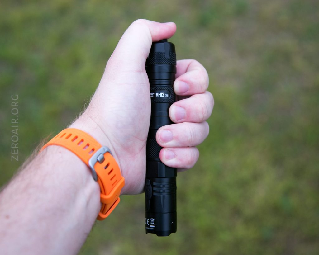 A person wearing an orange wristwatch holds a black flashlight in their right hand against a blurred outdoor background. The flashlight has visible text and details on its body.