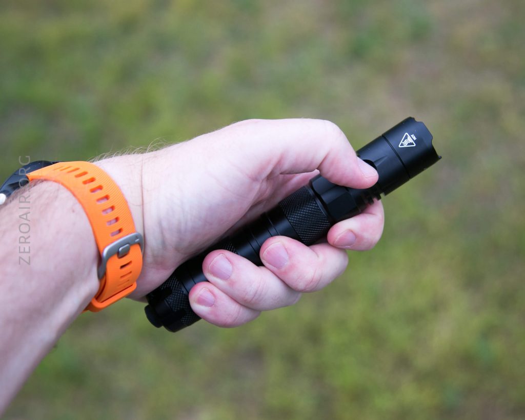 A person wearing an orange wristband holds a black flashlight in their right hand with a grassy background visible.