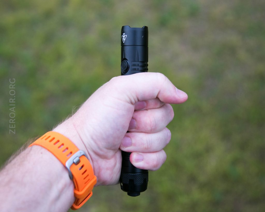 A person with a light skin tone wearing an orange wristwatch holds a black flashlight in their right hand against a blurred green and brown outdoor background.
