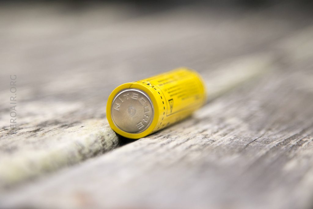 A yellow cylindrical battery labeled NITECORE lies on its side on a weathered wooden surface. The image focuses on the batterys metal end, with the label and background blurred.