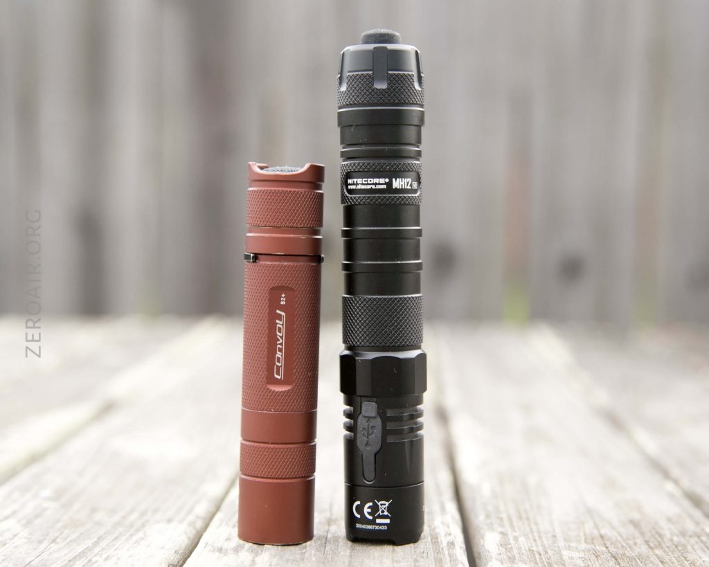 A small red flashlight and a larger black flashlight are standing upright side by side on a wooden surface with a blurred wooden fence in the background.