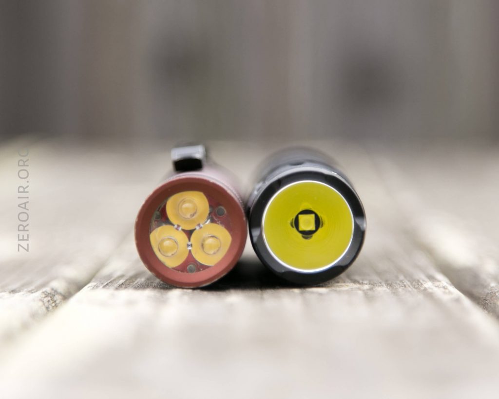 Two flashlights are shown lying side by side on a wooden surface, viewed from the front. The left flashlight has three LED bulbs, while the right flashlight has a single central LED bulb.