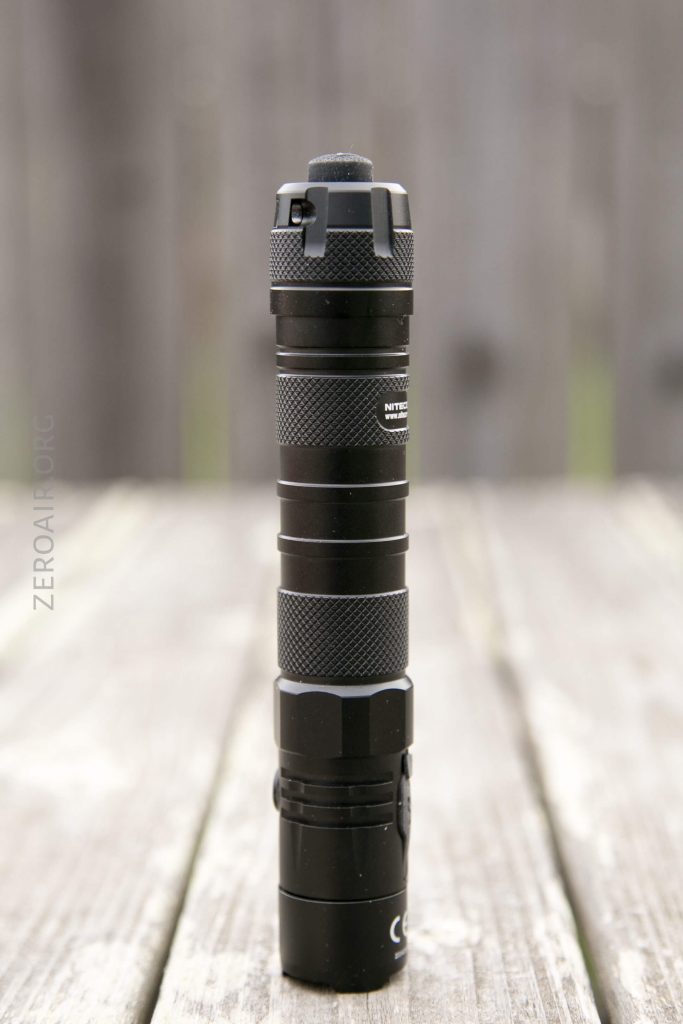 A black, cylindrical flashlight stands upright on a wooden surface with a blurred wooden fence in the background. The flashlight features a textured grip and a switch on the top.