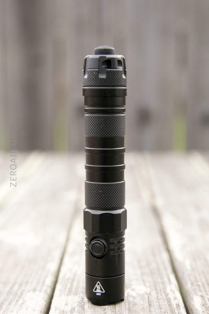 A black tactical flashlight stands upright on a wooden surface outdoors. The flashlight features a textured grip, side button, and warning symbol near the base. The background is blurred.