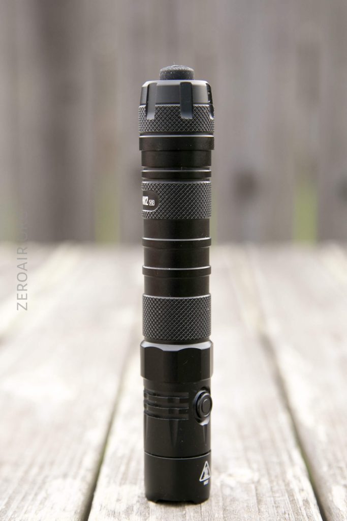 A black, cylindrical flashlight with a textured grip stands upright on a wooden surface, with a blurred, brownish background. The flashlight features a side button and a slightly raised tailcap switch.