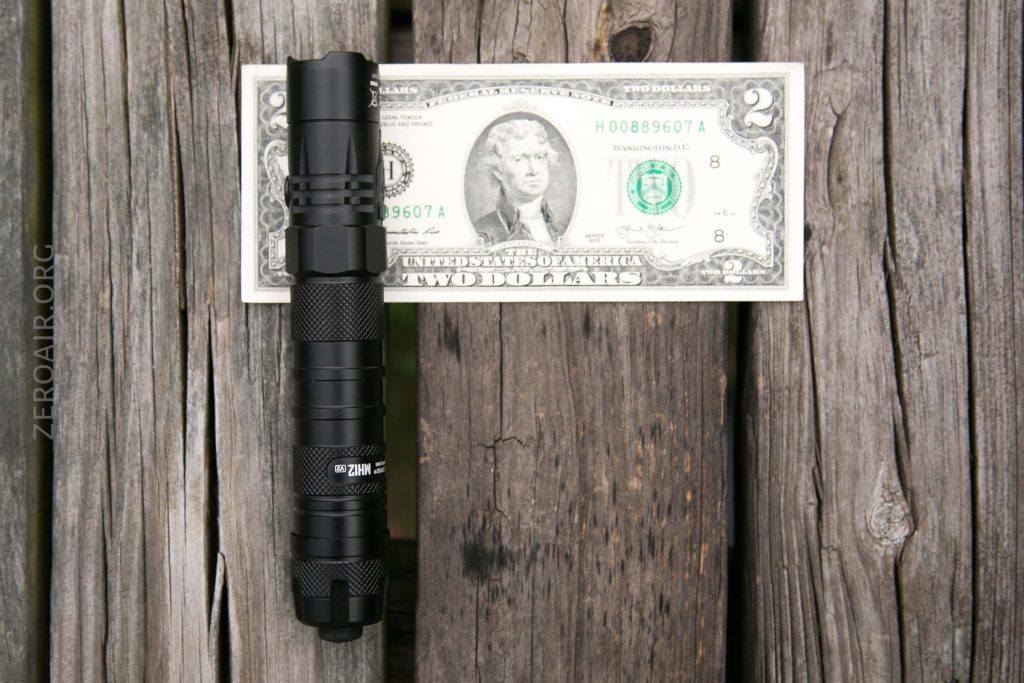 A black flashlight is placed vertically on a wooden surface next to a two-dollar bill laid flat, showing the relative size of the flashlight compared to the bill.