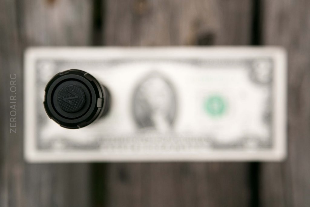 A close-up, top-down view of a black cylindrical object placed vertically on top of a blurred U.S. one dollar bill on a wooden surface. The text “ZEROAIR.ORG” is visible along the left side.