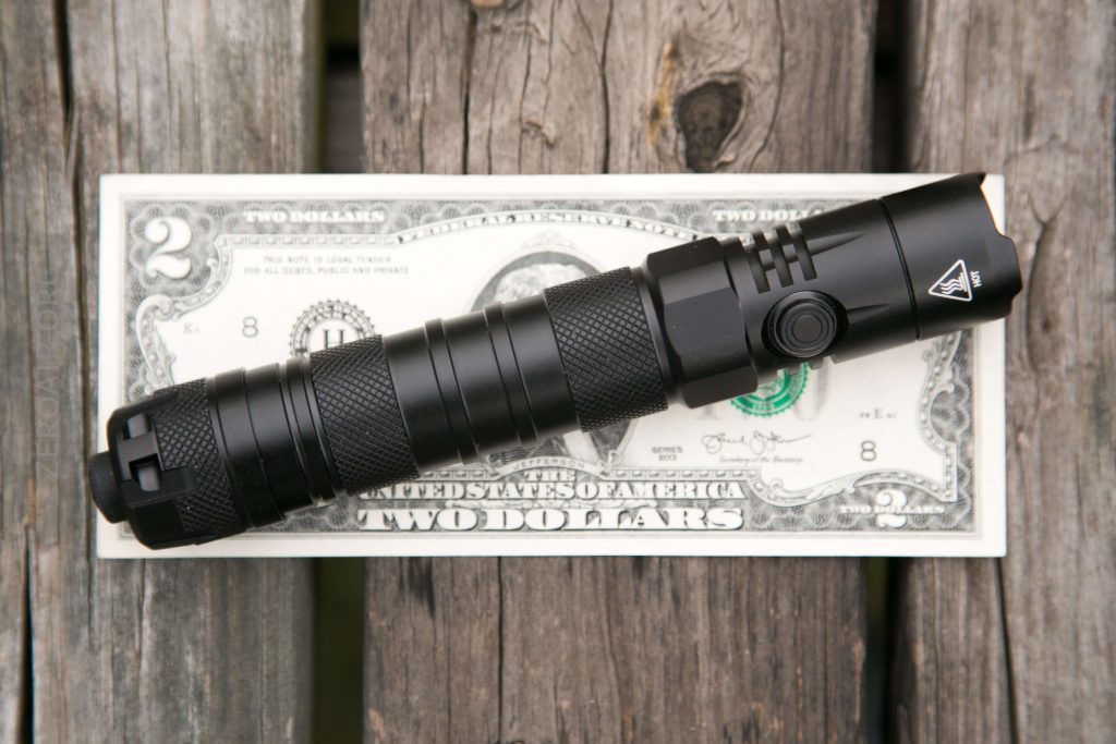 A black flashlight is placed horizontally on top of a two-dollar bill, which is lying on a weathered wooden surface.