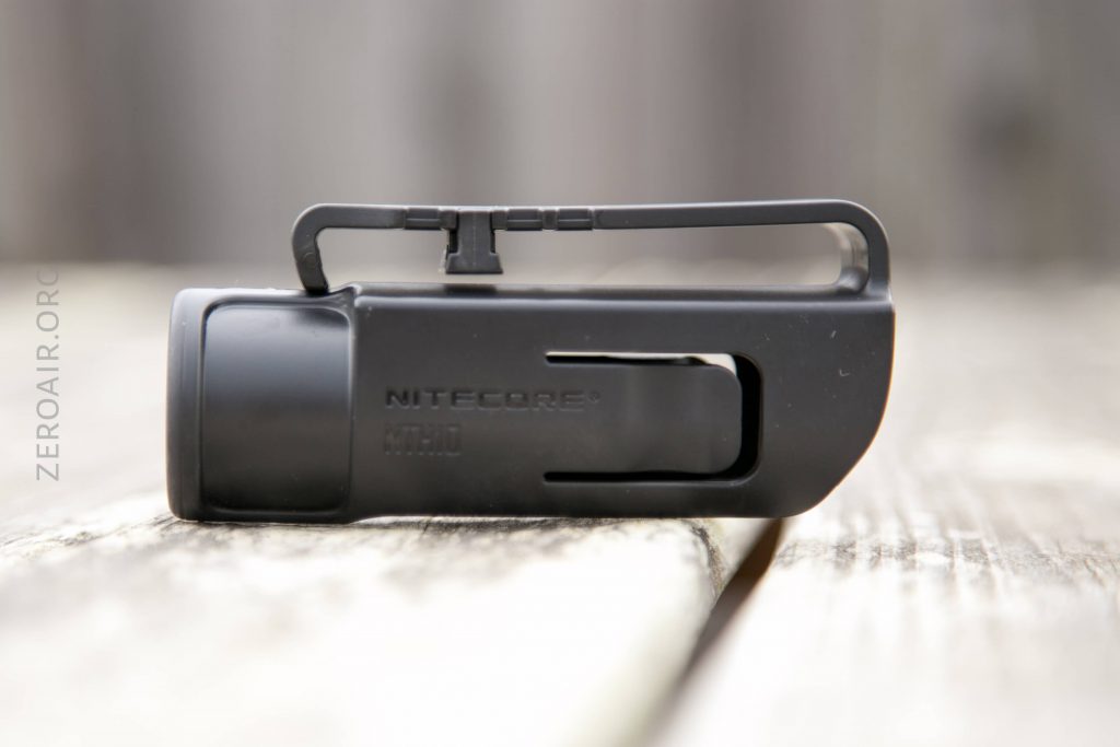 A close-up side view of a black Nitecore flashlight holster with a clip, placed on a weathered wooden surface. The background is blurred.