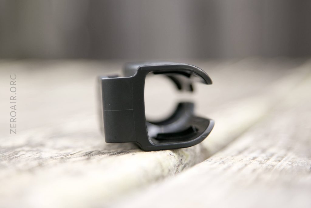 A small black plastic clip with an open, U-shaped design is resting on a wooden surface. The background is blurred and the word ZEROAIR.ORG is faintly visible on the left side.