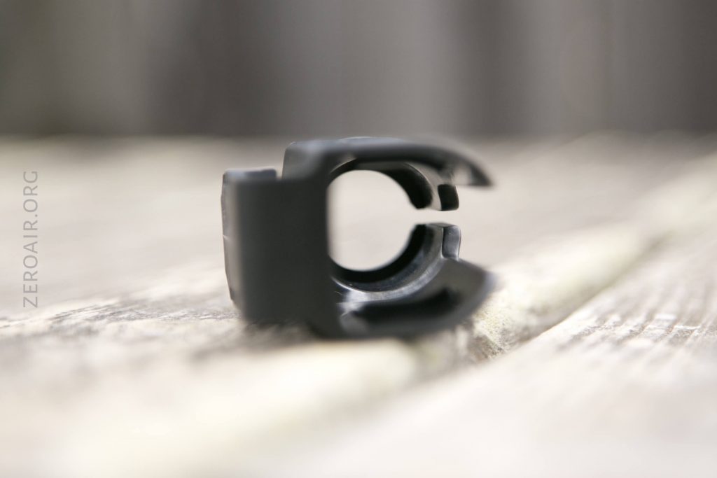 Close-up view of a black plastic or metal clip lying on its side on a wooden surface, with the background softly blurred. The text “ZEROAIR.ORG” is faintly visible on the left side of the image.