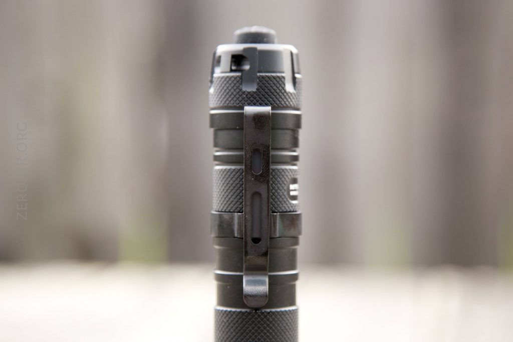 A close-up of a black textured flashlight standing upright on a blurred background, showing its metal clip and top button.