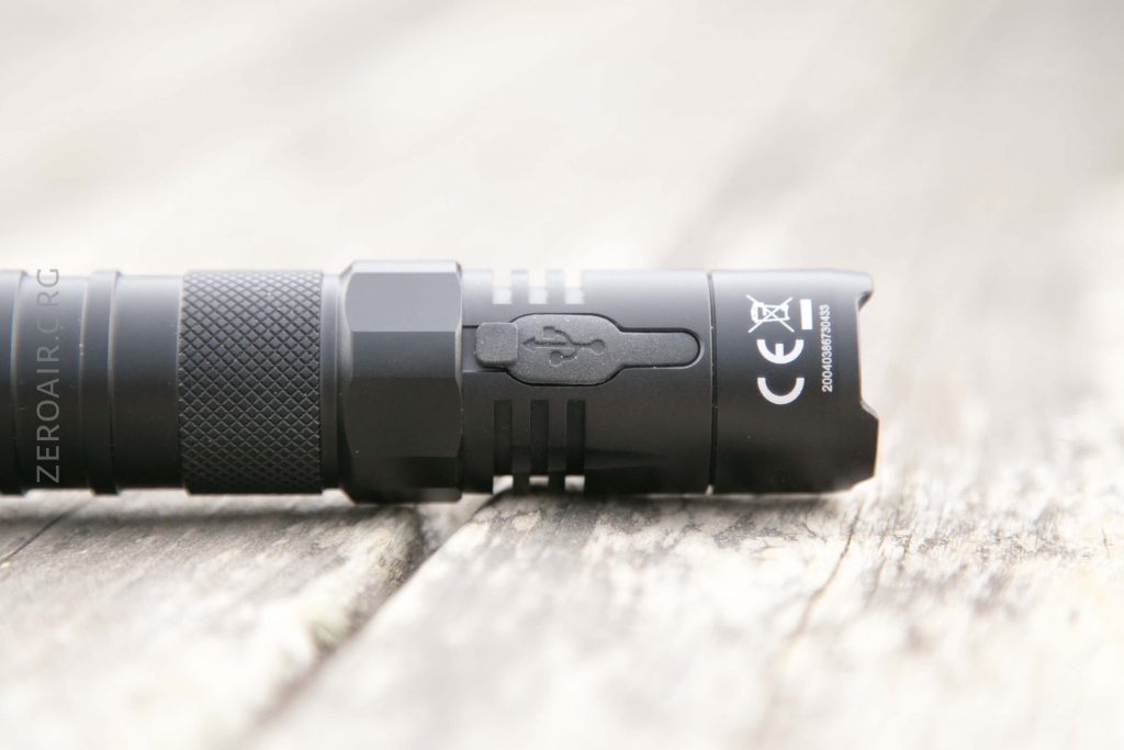 A black flashlight lying on its side on a wooden surface, showing a close-up of the body with a USB port covered by a rubber flap and certification markings.