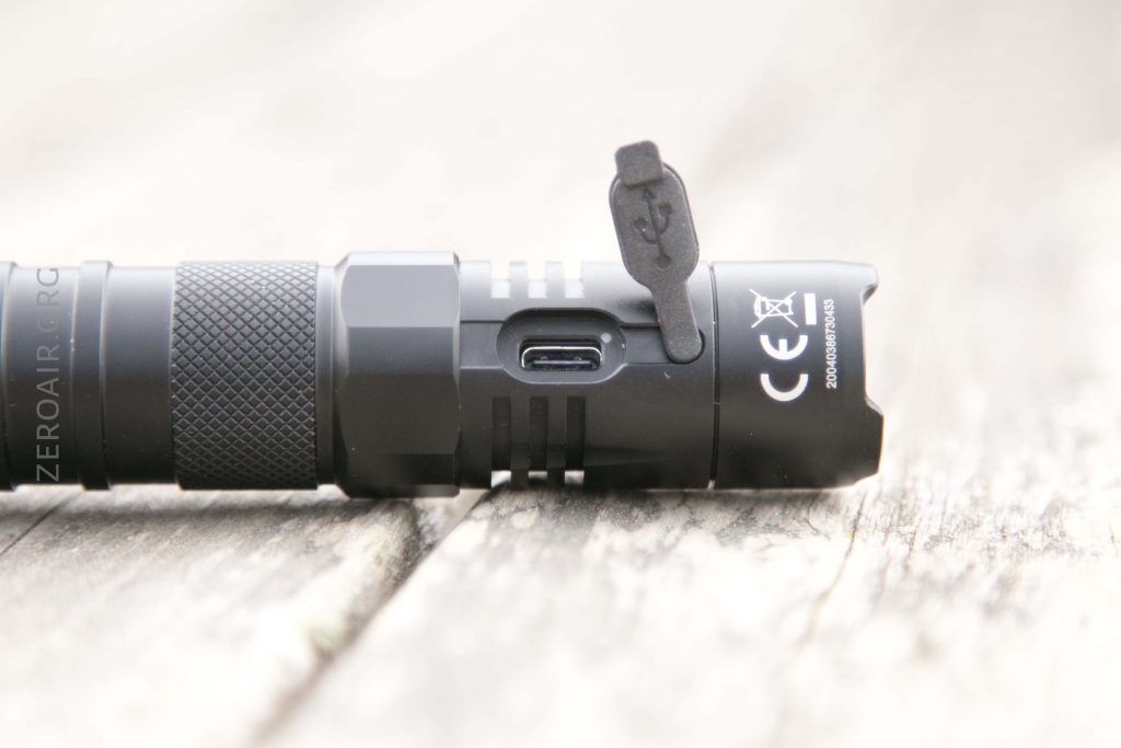A close-up view of a black flashlight lying on a wooden surface, showing its USB charging port with the rubber cover open and CE markings visible next to the port.