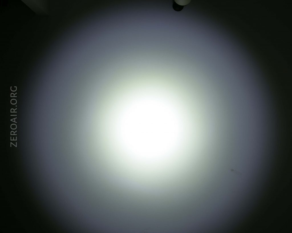 A bright, circular beam of white light is projected onto a plain wall, creating a gradient from white at the center to gray at the edges. The text “ZEROAIR.ORG” is visible along the left side.