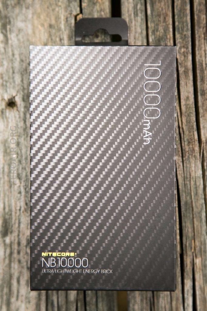 A black carbon-fiber textured box labeled 10000mAh and Nitecore NB10000 Ultralightweight Energy Brick sits on a weathered wooden surface.
