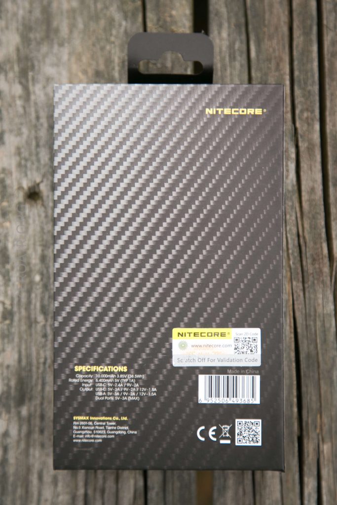 The back of a Nitecore product box with a carbon fiber pattern, specifications, QR codes, barcode, and a Scratch Off For Validation Code label visible; photographed on a weathered wooden surface.