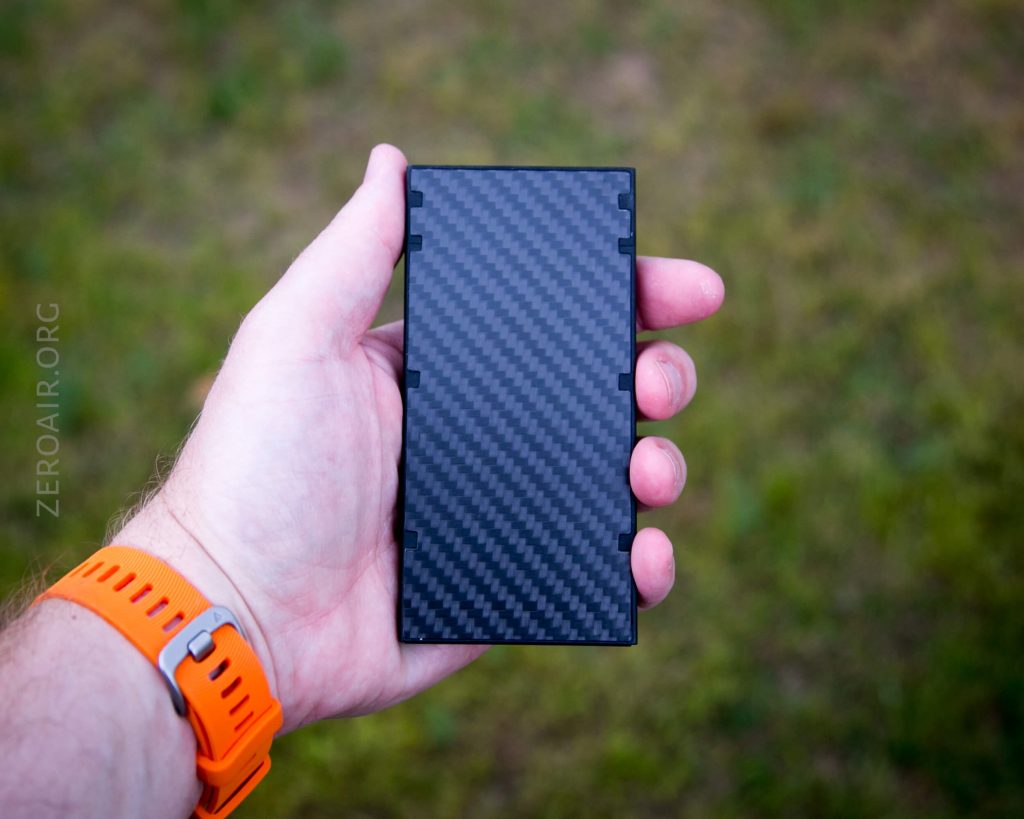 A person wearing an orange wristband holds a rectangular, black, carbon fiber-patterned object in their hand against a blurred grassy background. The word ZEROAIR.ORG is faintly visible on the left.