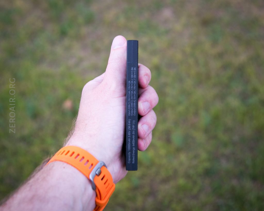 A person wearing an orange wristwatch holds a rectangular black battery with white text on its side against a blurred grassy background.
