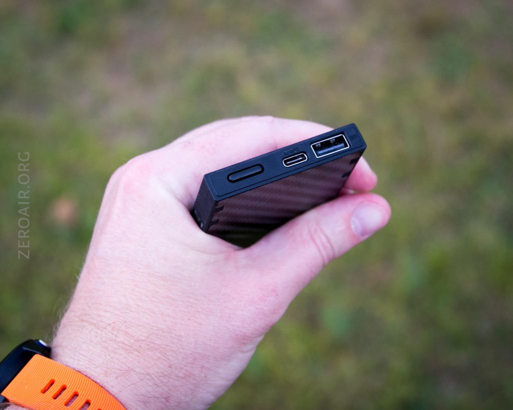 A hand holding a rectangular black electronic device with three ports: a micro USB, a USB-A, and a smaller slot. The background is grassy and blurred. The person is wearing an orange wristband.