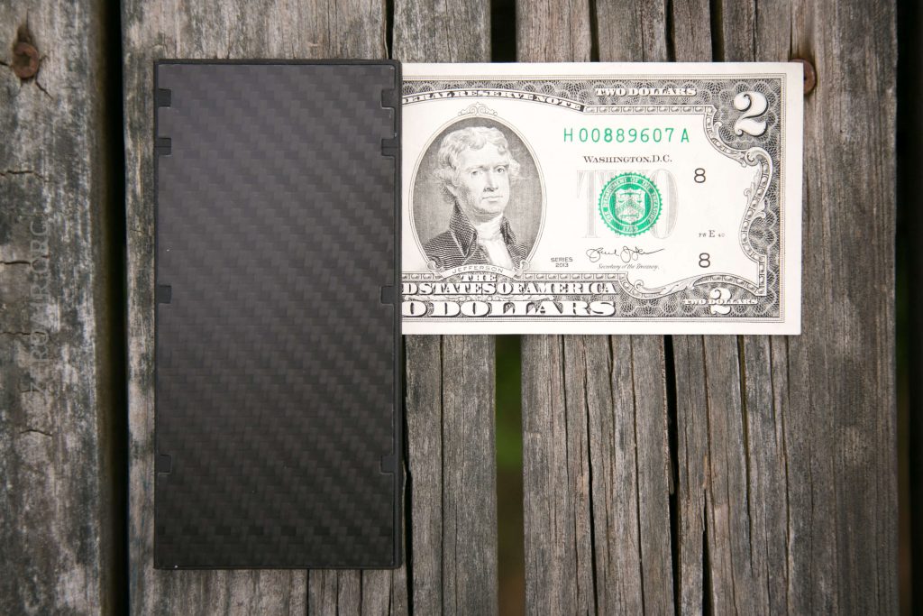 A rectangular black object with a textured pattern is placed next to a two-dollar bill on a weathered wooden surface. The objects are shown side by side for size comparison.