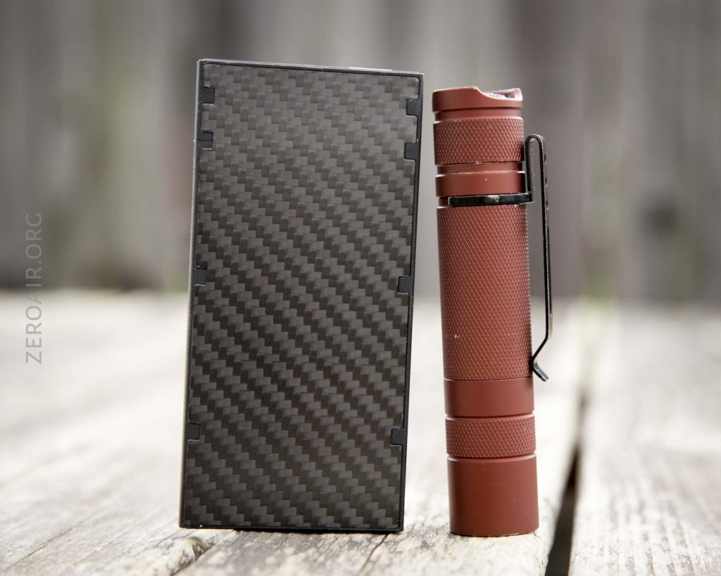 A rectangular black carbon-fiber patterned object is placed next to a small, textured, reddish-brown flashlight with a black clip, both standing upright on a wooden surface.
