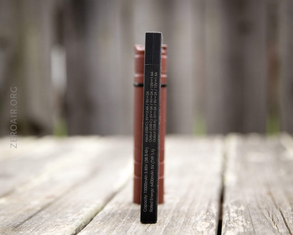 A close-up of a black rectangular battery charger standing upright on a wooden surface outdoors, with blurred cylindrical batteries and a wooden fence in the background. White text is printed on the side of the charger.