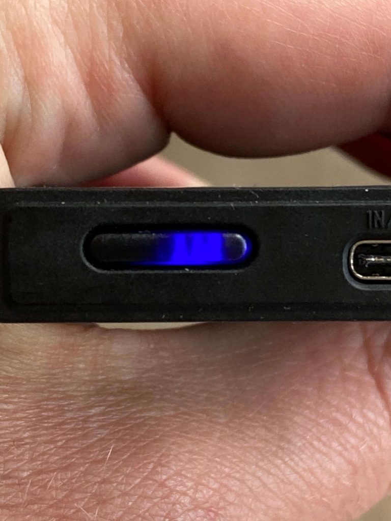 A close-up view of a hand holding a black electronic device with a rectangular blue LED indicator illuminated and a USB-C port visible on the right side.