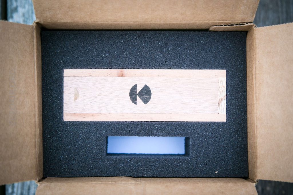 A rectangular wooden block with a black geometric logo rests in a black foam insert inside an open cardboard box. The foam has a rectangular cutout below the block.