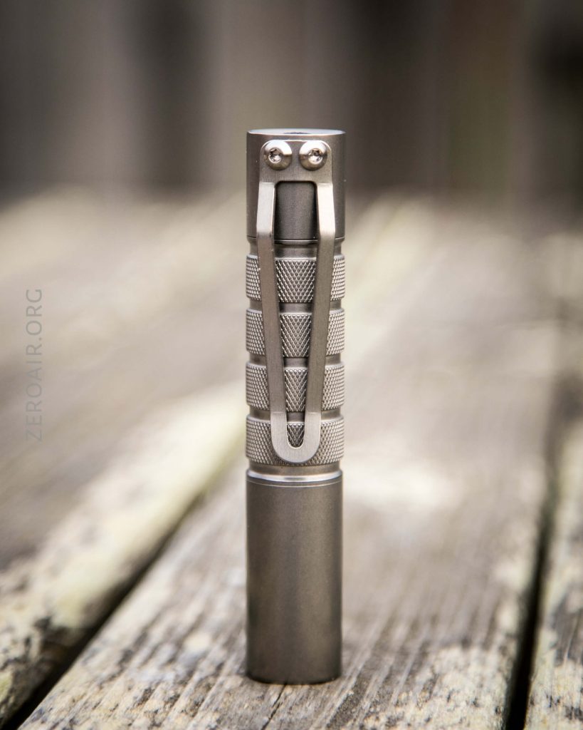 A small, metallic flashlight stands upright on a wooden surface, featuring a textured grip and a metal pocket clip.