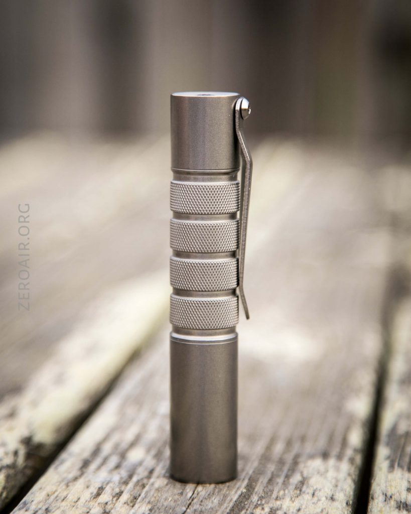 A small, metallic flashlight with a textured grip and a pocket clip stands upright on a weathered wooden surface. The background is blurred.