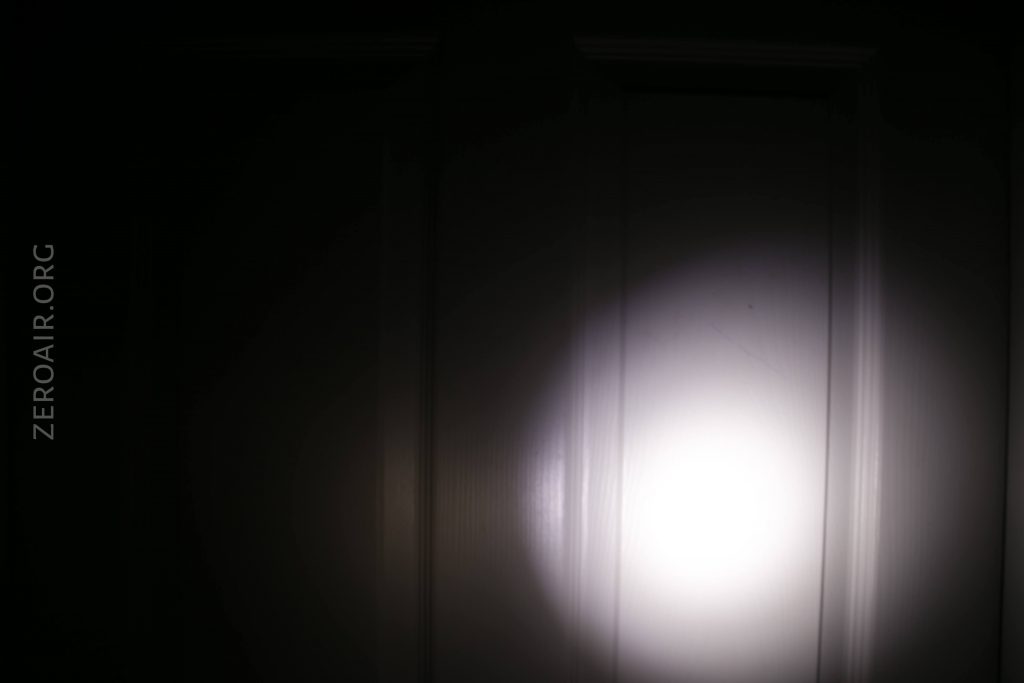 A bright flashlight beam shines on a dark, paneled door. The center of the light is intense, fading outward. The text ZEROAIR.ORG is visible vertically along the left side of the image.