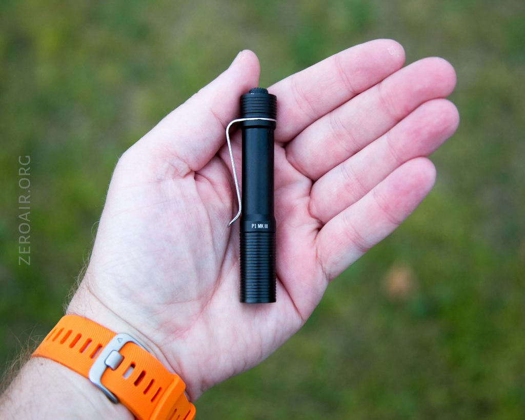 A hand with a light skin tone and an orange wristwatch holds a small black flashlight with a pocket clip against a blurred outdoor background.