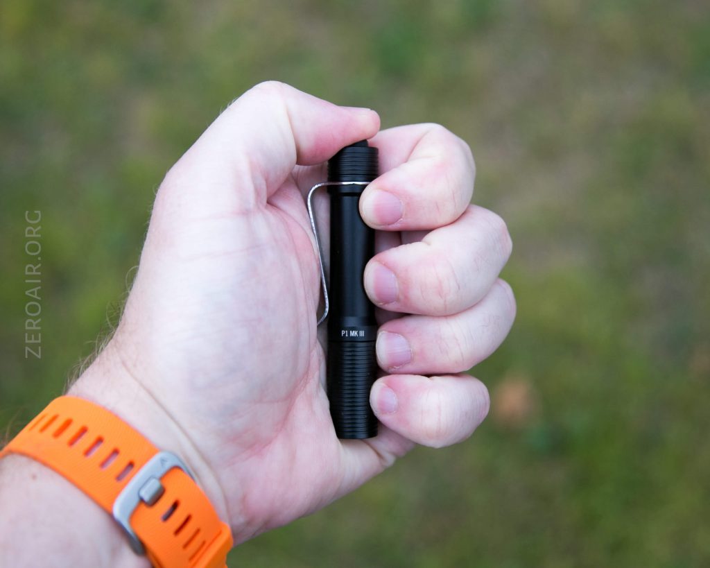 A person wearing an orange wristband holds a small black flashlight with a clip in their right hand against a blurred green background. The flashlight is marked PL Mini.