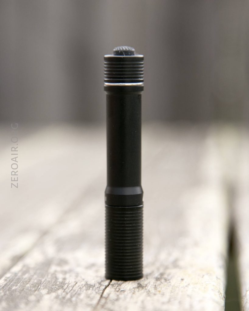 A black, cylindrical flashlight standing upright on a weathered wooden surface with a blurred outdoor background.
