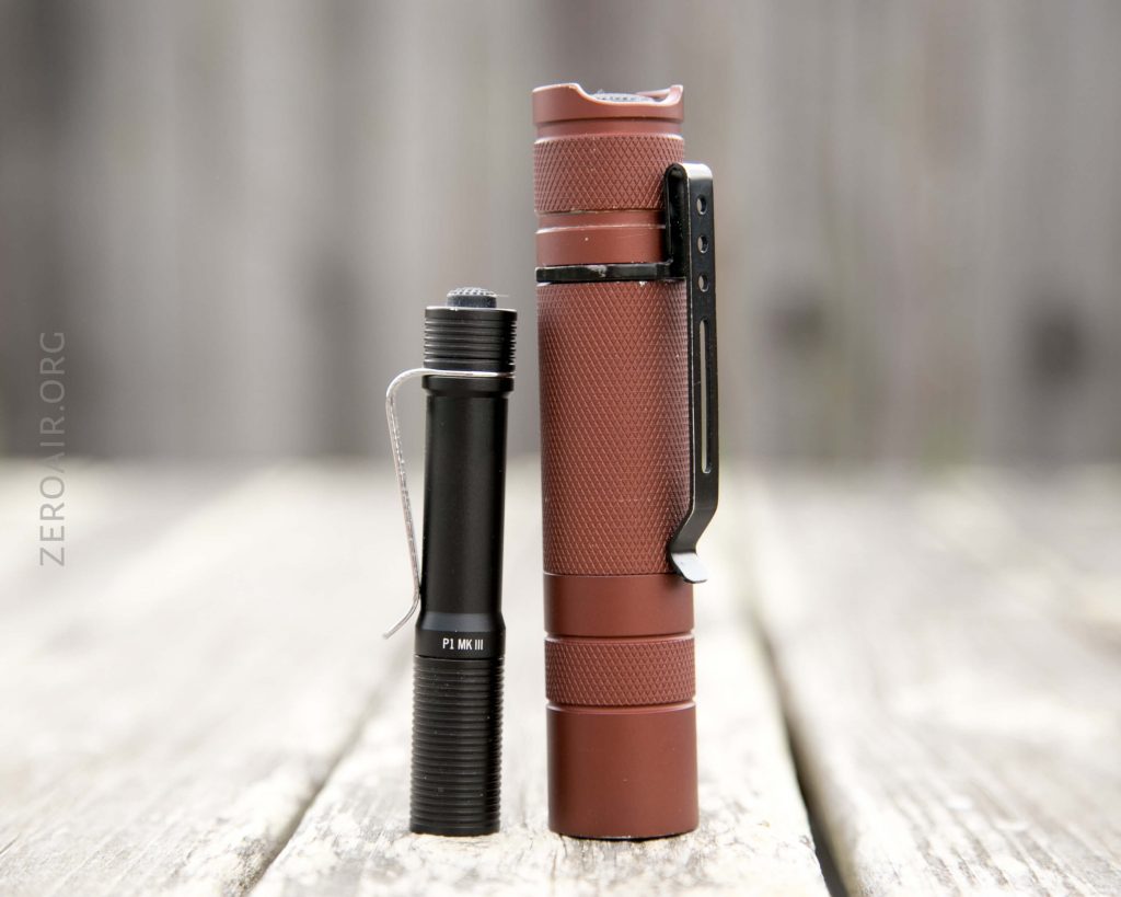 A small black flashlight and a larger brown flashlight stand upright side by side on a wooden surface with a blurred background. Both have pocket clips.