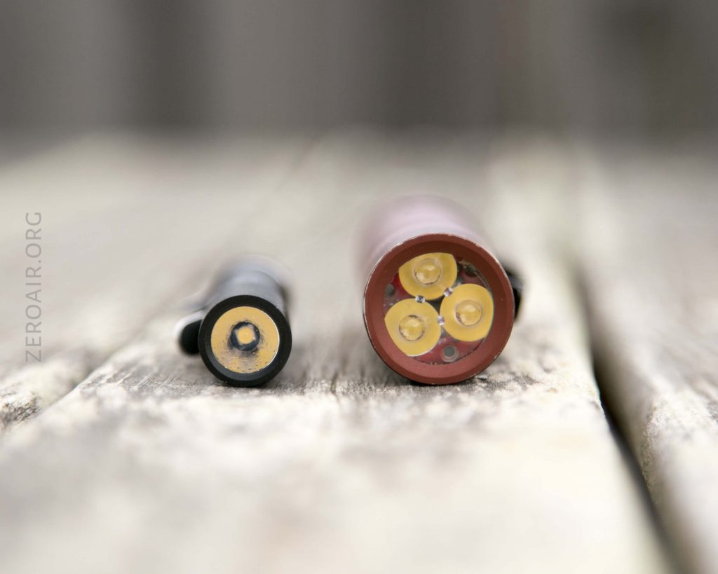Two cylindrical flashlight components lie on a wooden surface. The left object is smaller with a single central contact, while the right object is larger and shows four yellow LED emitters arranged in a circle.