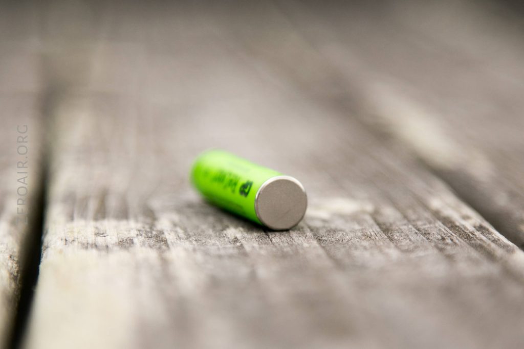 A green cylindrical battery lying on its side on a weathered wooden surface, with the focus on the metal end of the battery and the background slightly blurred.