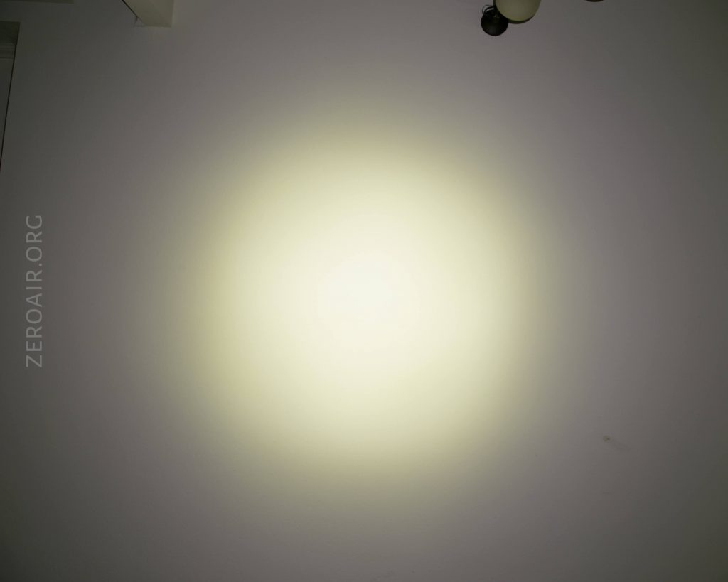 A circular beam of yellowish light is projected onto a plain white wall. The words ZEROAIR.ORG appear vertically along the left side. A ceiling fixture is partially visible in the top right corner.