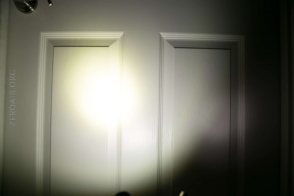 A bright flashlight beam illuminates the center of a white paneled door, with the edges of the door fading into shadow. The text ZEROAIR.ORG is visible on the left side of the image.